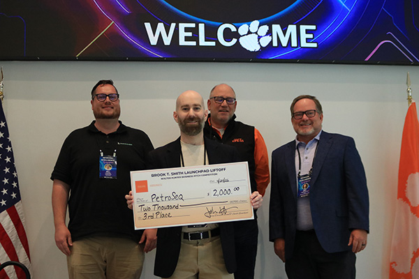 Award recipient and judges holding oversized checks beneath a large welcome banner with U.S. and Clemson flags beside them.