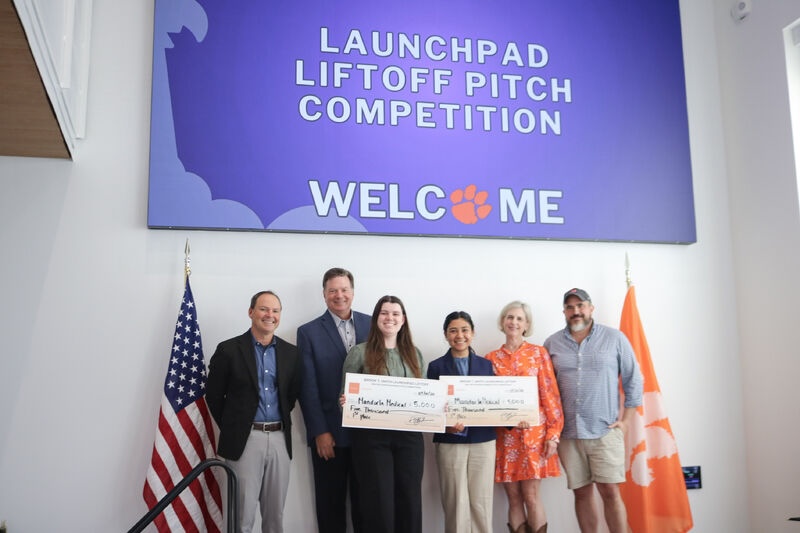 Group of award recipients and organizers holding oversized checks beneath a large “Launchpad Liftoff Pitch Competition” welcome banner, with U.S. and Clemson flags beside them.