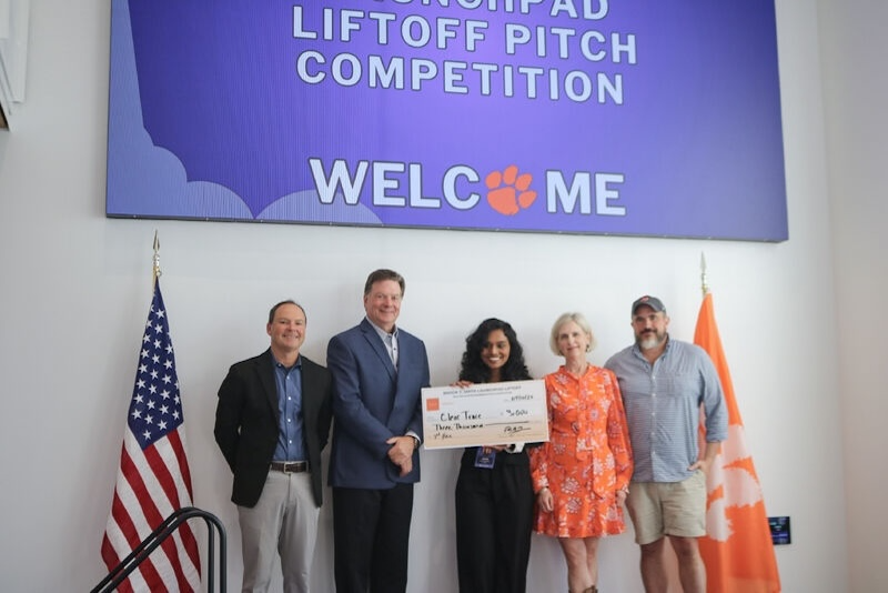 Five people pose on a stage holding an oversized check beneath a large screen that reads “Liftoff Pitch Competition” and “Welcome,” with U.S. and Clemson flags on either side.