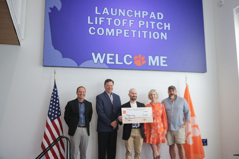 Group of five people on a stage holding a large check beneath a screen that reads “Launchpad Liftoff Pitch Competition” and “Welcome,” with U.S. and Clemson flags beside them.