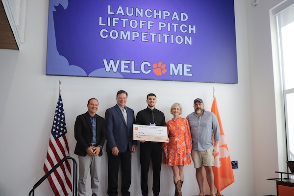 Group of five people posing indoors beneath a “Launchpad Liftoff Pitch Competition” welcome sign, with one person holding an oversized check and U.S. and orange flags on either side.