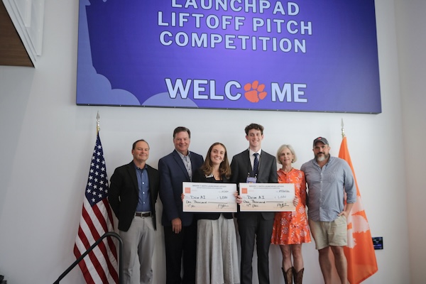 Group of people on a stage holding oversized checks under a “Launchpad Liftoff Pitch Competition” welcome banner, with U.S. and Clemson flags on either side.
