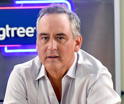 Close-up of a middle-aged man in a light button-down shirt, seated indoors with a blurred neon sign in the background.
