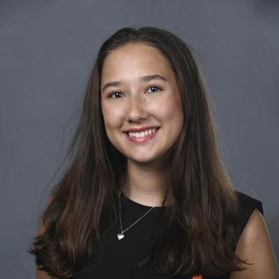 Studio headshot of a smiling young woman with long dark hair against a gray background.