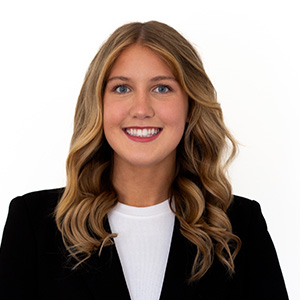 Headshot of a smiling woman with long wavy hair, wearing a black blazer over a white top against a white background.