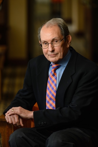 Portrait of a distinguished older man in a suit and colorful tie, seated in a warm-toned interior.