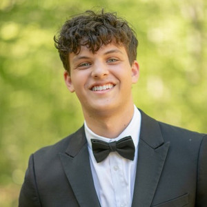 A smiling young man in a tuxedo with a bow tie stands against a blurred green background.