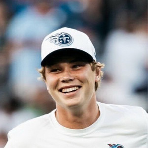 Smiling young man wearing a white cap with a logo, likely at a sporting event.