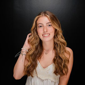 A portrait of a young woman with long, wavy hair and a warm smile, standing against a dark backdrop.