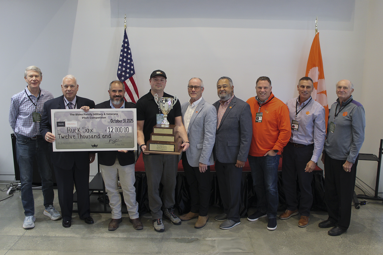 A group of individuals stands on stage holding a large check and a trophy, celebrating a donation of $12,000 to Ruck Sox, flanked by American and Clemson University flags.