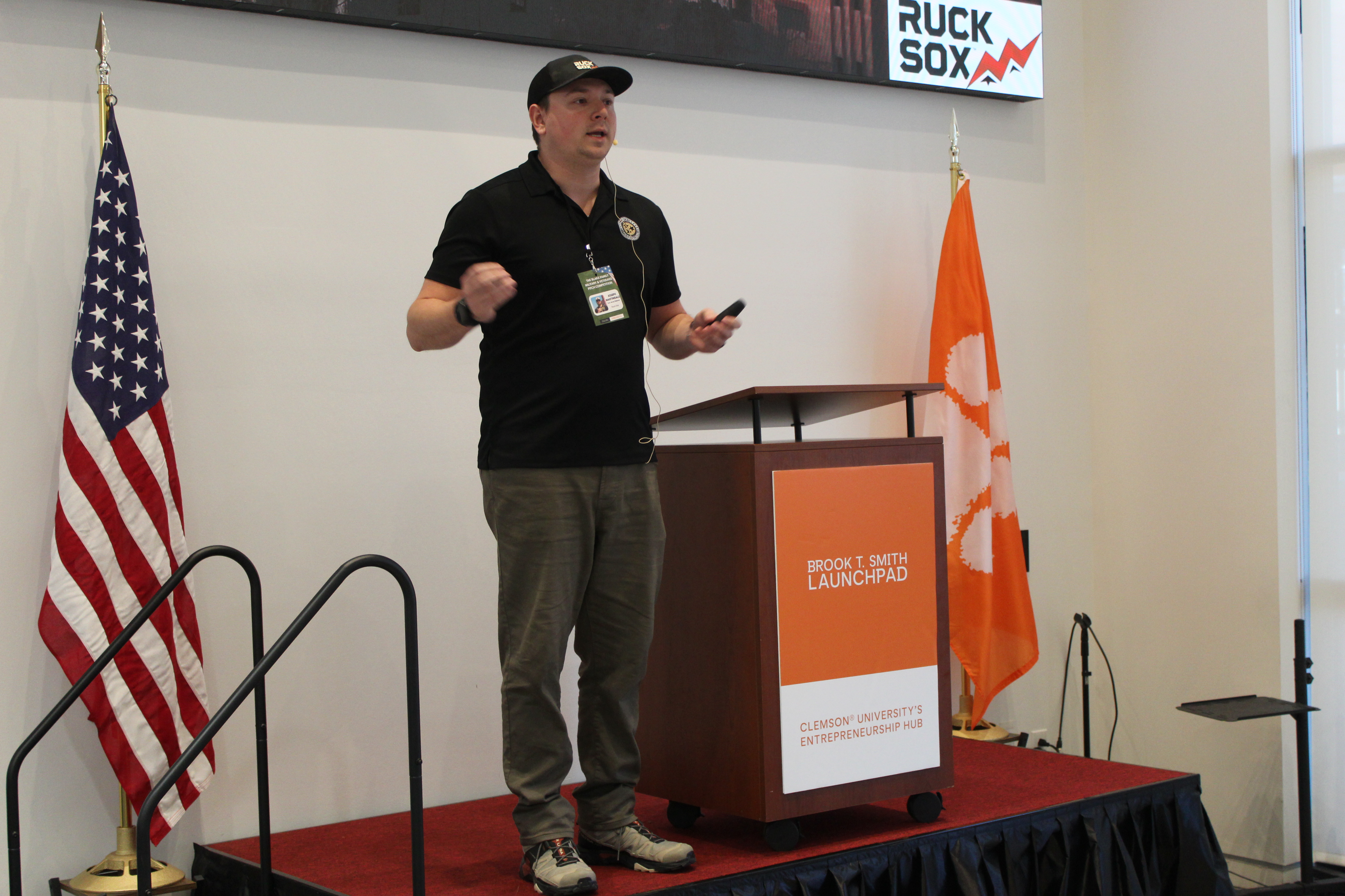 Speaker giving a presentation on stage next to a podium at Clemson University’s Brook T. Smith Launchpad, with U.S. and Clemson flags behind him.