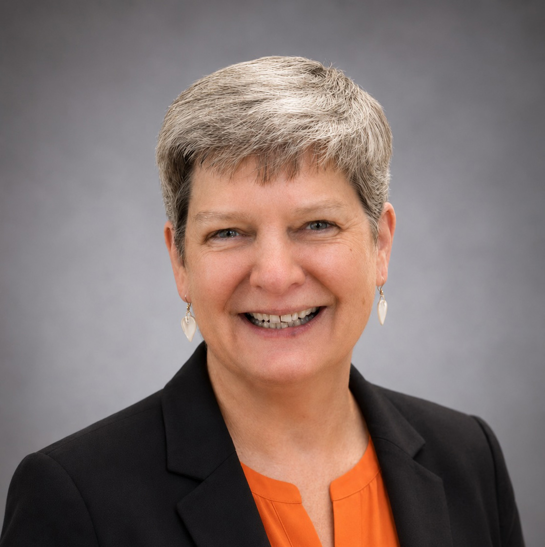 Smiling person with short gray hair wearing a black blazer and orange top against a neutral gray background.
