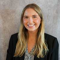 Professional headshot of a smiling woman with long blonde hair wearing a black blazer against a gray studio backdrop.