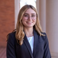 Smiling young woman wearing glasses and a dark blazer, standing outdoors near a column.