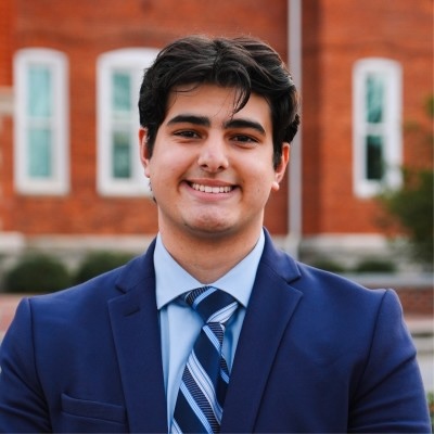 Smiling person in a navy suit and striped tie standing outdoors in front of a brick building.