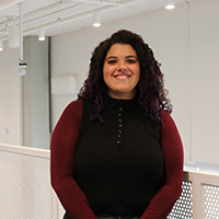 Smiling person with curly hair wearing a black top with burgundy sleeves, standing indoors by a railing.