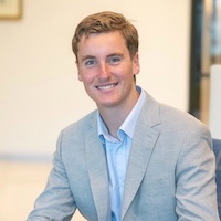 Smiling man in a light gray blazer sitting indoors.