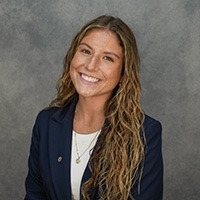 Smiling woman with long wavy hair wearing a navy blazer and white top against a gray studio backdrop.