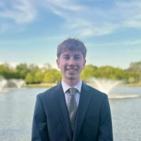 Young man in a suit and tie smiling in front of a lake with fountains.