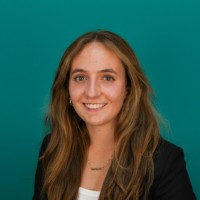 Smiling woman with long brown hair in a black blazer against a teal background.