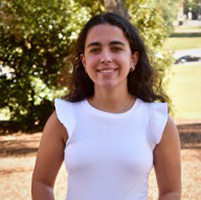 Smiling person in a white sleeveless top standing outdoors with trees in the background.