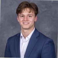 Portrait of a young man in a blue suit jacket and white shirt, smiling against a gray studio background.