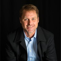 Professional portrait of a smiling man wearing a black coat and light blue shirt, set against a dark background.