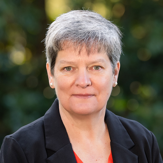 Headshot of a short-haired person wearing a dark blazer, photographed outdoors with a blurred green background.