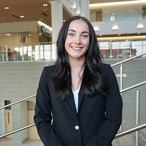 Smiling woman in a black blazer standing indoors in a modern building with railings and large windows in the background.