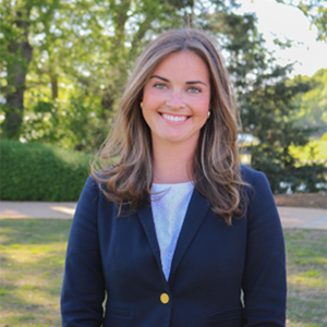 A smiling woman in a blazer stands outdoors, surrounded by greenery.