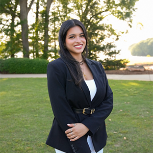 A young woman in a black blazer standing outdoors in a sunny setting, smiling confidently.