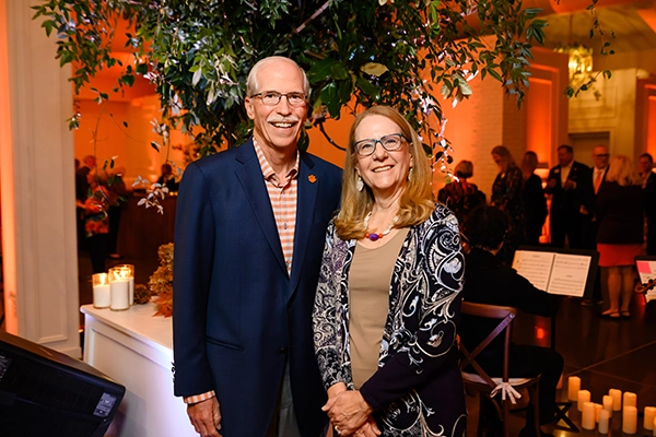A smiling man and woman pose together at an indoor event with warm orange lighting, candles, and other guests in the background.