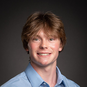 Headshot of a young man with light brown hair wearing a blue shirt, smiling against a dark background.