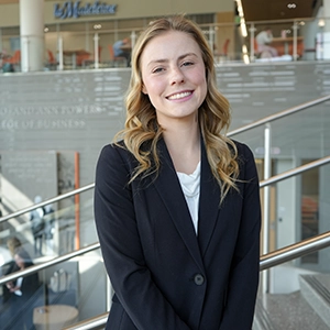 Smiling woman in a dark blazer stands indoors near a railing in a modern building lobby.