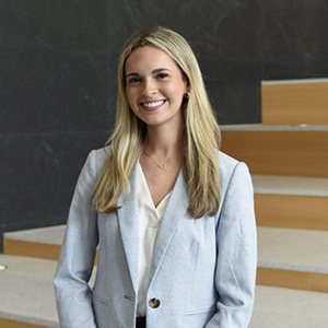 A smiling woman with long, wavy hair wearing a light blazer and a white blouse, standing on a set of wooden steps.