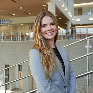 Smiling woman in a gray blazer standing indoors by a metal railing, with a bright modern atrium in the background.