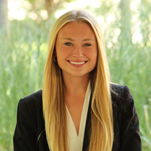 Professional portrait of a young woman with long blonde hair, wearing a black blazer and white top, set against a backdrop of greenery.