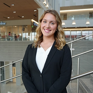 Smiling woman in a black blazer and white blouse standing indoors in a modern office atrium with railings and glass in the background.