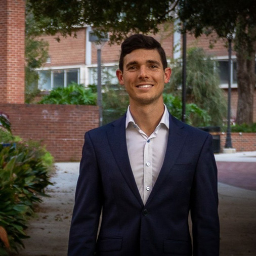 A smiling man in a suit stands confidently outdoors, surrounded by greenery and brick architecture.