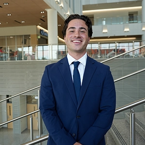 Smiling man in a blue suit and tie standing in a modern building atrium with stairs and railings in the background.