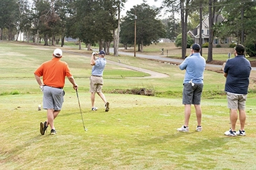 A group of four men watching a golfer take a swing on a golf course.