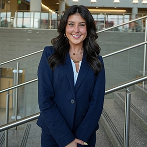 Smiling woman in a navy suit standing indoors by a metal railing in a modern building.