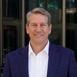 Headshot of a smiling man in a suit, standing outside a modern building.
