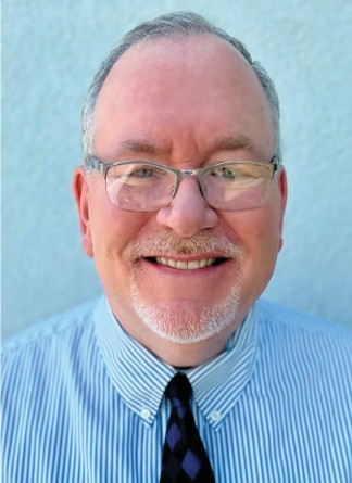 Smiling man with glasses wearing a striped dress shirt and tie against a light blue background.