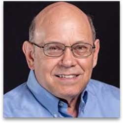 Smiling older man wearing glasses and a light blue collared shirt against a dark background.