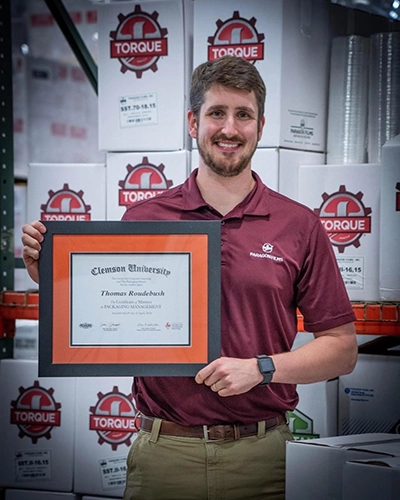 Man holding a framed Clemson University certificate while standing in a warehouse with stacked Torque-branded boxes behind him.