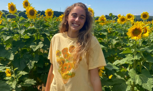 A blonde young woman smiles in front of sunflowers.