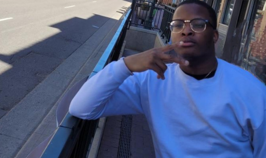 A young man in a white shirt holds up a peace sign in front of a street of buildings.