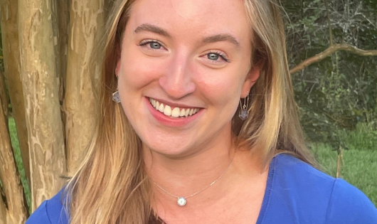 A blonde young woman in a blue shirt smiles at the camera.