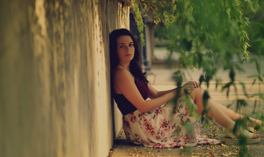 A young woman with brown hair sits in the shade by a concrete wall.